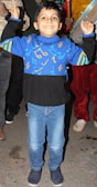 A young child is standing with raised arms, smiling brightly. They are wearing a vibrant blue sweater adorned with colorful musical notes and symbols, paired with light blue jeans and navy blue shoes. The background includes adults, with one holding a musical instrument.