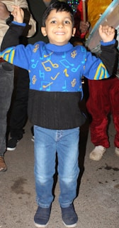 A young child is standing with raised arms, smiling brightly. They are wearing a vibrant blue sweater adorned with colorful musical notes and symbols, paired with light blue jeans and navy blue shoes. The background includes adults, with one holding a musical instrument.