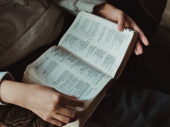 A person is holding an open book with text printed in an East Asian script, likely reading or studying the material. The book appears to be well-used, with slightly worn edges. The person is wearing casual clothing, sitting comfortably as they engage with the text.