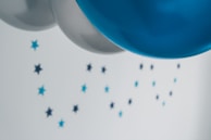 Several balloons in shades of silver and blue float in the foreground, while small star-shaped decorations, also in blue hues, are scattered across a light background.