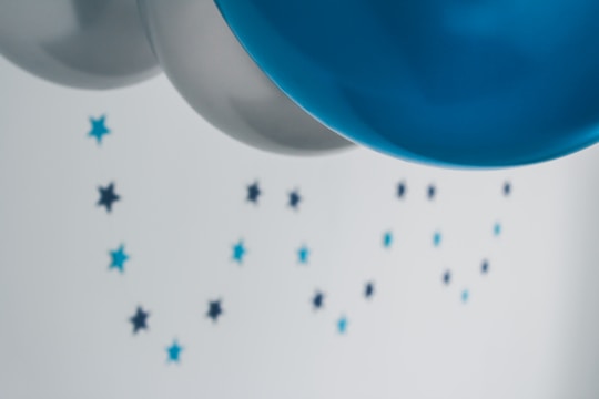 Several balloons in shades of silver and blue float in the foreground, while small star-shaped decorations, also in blue hues, are scattered across a light background.
