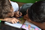 Two children are closely engaged with a book, lying on their sides. One child is pointing at a colorful illustration while the other writes or draws with a pen. The book appears to be open on a table, featuring large, vibrant illustrations, likely from a story or educational material.