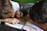Two children are closely engaged with a book, lying on their sides. One child is pointing at a colorful illustration while the other writes or draws with a pen. The book appears to be open on a table, featuring large, vibrant illustrations, likely from a story or educational material.