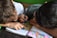 Two children are closely engaged with a book, lying on their sides. One child is pointing at a colorful illustration while the other writes or draws with a pen. The book appears to be open on a table, featuring large, vibrant illustrations, likely from a story or educational material.
