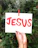 A hand with dark skin tone holding a white card with the word 'JESUS' painted in bold red letters. The card is clipped to a green plant background using a red clothespin.