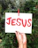 A hand with dark skin tone holding a white card with the word 'JESUS' painted in bold red letters. The card is clipped to a green plant background using a red clothespin.
