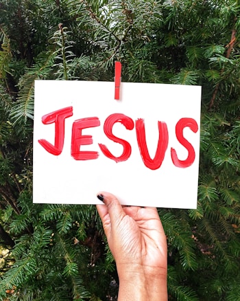 A hand with dark skin tone holding a white card with the word 'JESUS' painted in bold red letters. The card is clipped to a green plant background using a red clothespin.
