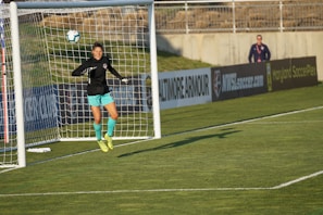 A soccer player, dressed in a black jersey and turquoise shorts, jumps in mid-air to make a save near a goal post. The player is wearing fluorescent yellow cleats. In the background, a person stands by the advertisement boards lining the edge of the soccer field.