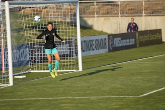 A soccer player, dressed in a black jersey and turquoise shorts, jumps in mid-air to make a save near a goal post. The player is wearing fluorescent yellow cleats. In the background, a person stands by the advertisement boards lining the edge of the soccer field.