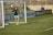 A soccer player, dressed in a black jersey and turquoise shorts, jumps in mid-air to make a save near a goal post. The player is wearing fluorescent yellow cleats. In the background, a person stands by the advertisement boards lining the edge of the soccer field.