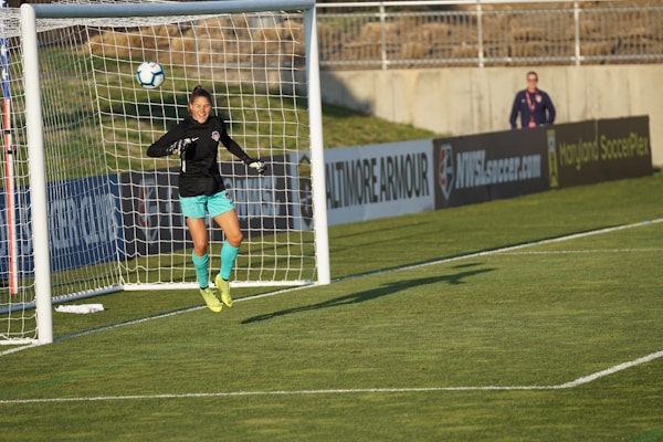 A soccer player, dressed in a black jersey and turquoise shorts, jumps in mid-air to make a save near a goal post. The player is wearing fluorescent yellow cleats. In the background, a person stands by the advertisement boards lining the edge of the soccer field.