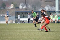 Two soccer players are actively competing for the ball on a grassy field, with a referee and additional players in the background. The player in red is wearing number 28 and is being closely challenged by an opponent in a beige uniform. Other team members and coaches are visible on the sidelines, observing the match.