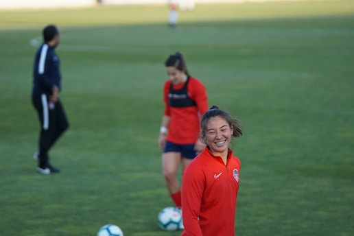 Players are on a grassy sports field, focusing on a woman in a red sports jersey smiling. Other individuals are present, wearing similar sports attire. Soccer balls are on the ground.