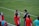A group of people wearing sports attire are gathered on a grassy field. Some individuals are dressed in red jackets, while others are in turquoise and navy blue. A coach appears to be giving instructions. Sports equipment, such as water bottles and a bench with towels, can be seen nearby.