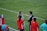 A group of people wearing sports attire are gathered on a grassy field. Some individuals are dressed in red jackets, while others are in turquoise and navy blue. A coach appears to be giving instructions. Sports equipment, such as water bottles and a bench with towels, can be seen nearby.