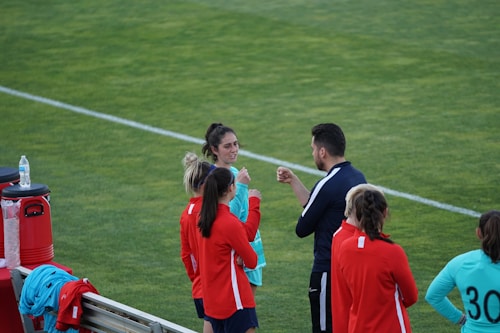 A group of people wearing sports attire are gathered on a grassy field. Some individuals are dressed in red jackets, while others are in turquoise and navy blue. A coach appears to be giving instructions. Sports equipment, such as water bottles and a bench with towels, can be seen nearby.