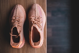 A pair of stylish, knitted sneakers in a peachy color are placed side by side on a wooden surface. The footwear features intricate patterns and laces, suggesting a modern and trendy design.