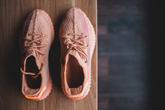 A pair of stylish, knitted sneakers in a peachy color are placed side by side on a wooden surface. The footwear features intricate patterns and laces, suggesting a modern and trendy design.