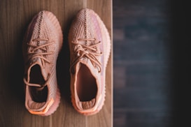 A pair of stylish, knitted sneakers in a peachy color are placed side by side on a wooden surface. The footwear features intricate patterns and laces, suggesting a modern and trendy design.