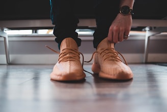 A close-up view of a person tying the laces of a stylish, beige sneaker. The person is wearing a black watch and a ring on the left hand, while the floor is made of polished wood. The scene appears casual and focused on the action of tying shoelaces.