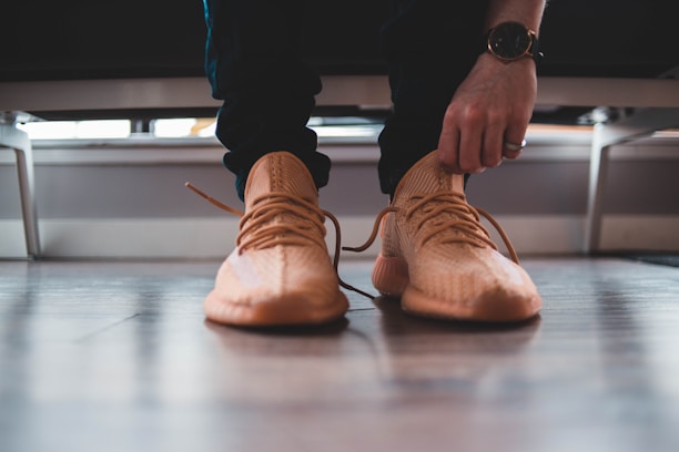 A close-up view of a person tying the laces of a stylish, beige sneaker. The person is wearing a black watch and a ring on the left hand, while the floor is made of polished wood. The scene appears casual and focused on the action of tying shoelaces.