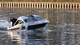 A small motorboat is moving swiftly across a body of water. The boat has a white and dark-colored hull, and a person is seated at the back, adjusting something on the boat. The water is reflecting the sunlight, creating a shimmering effect. In the background, there is a wooden or concrete barrier along the shoreline.