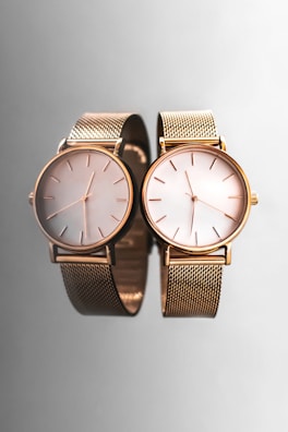 Two elegant wristwatches with gold mesh bands and minimalist dials are placed side by side against a smooth, reflective gray surface, creating a sophisticated and luxurious appearance.