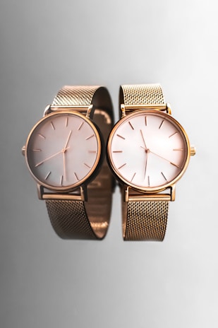 Two elegant wristwatches with gold mesh bands and minimalist dials are placed side by side against a smooth, reflective gray surface, creating a sophisticated and luxurious appearance.