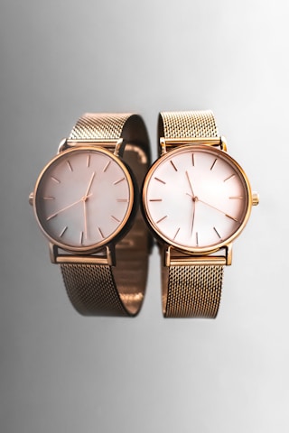 Two elegant wristwatches with gold mesh bands and minimalist dials are placed side by side against a smooth, reflective gray surface, creating a sophisticated and luxurious appearance.