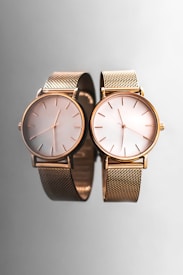 Two elegant wristwatches with gold mesh bands and minimalist dials are placed side by side against a smooth, reflective gray surface, creating a sophisticated and luxurious appearance.