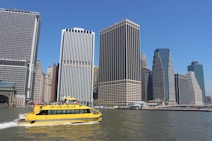 A bright yellow water taxi navigates through a body of water in front of a collection of skyscrapers forming a cityscape. The buildings, with their various architectural designs, rise against a clear blue sky, providing a stark contrast to the bustling water vessel.