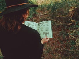 A person wearing a dark hat is holding an open book with botanical illustrations while standing amidst dense greenery. The book's pages feature detailed drawings of different plants. The surrounding area is filled with various types of plant life, creating a natural and serene setting.