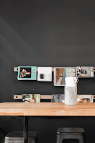 A modern interior scene featuring a black wall with a simple shelf displaying various magazines. The shelf is made of wood and mounted horizontally. Below the shelf, a wooden table with a vase or container holding napkins and straws is placed. Two metallic stools are positioned under the table, adding a touch of industrial style.