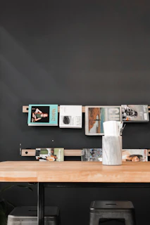 A modern interior scene featuring a black wall with a simple shelf displaying various magazines. The shelf is made of wood and mounted horizontally. Below the shelf, a wooden table with a vase or container holding napkins and straws is placed. Two metallic stools are positioned under the table, adding a touch of industrial style.