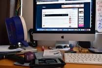 A workspace with an iMac displaying a website, surrounded by various office items including a keyboard, a small globe, a fan, papers, pens, and a pair of glasses. The desk also houses ornaments and electronic accessories.
