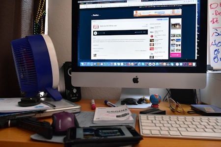 A workspace with an iMac displaying a website, surrounded by various office items including a keyboard, a small globe, a fan, papers, pens, and a pair of glasses. The desk also houses ornaments and electronic accessories.
