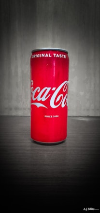 A tall, red soda can with white lettering stands on a flat surface, with a textured gray background. The can is prominently branded with the logo and slogan.