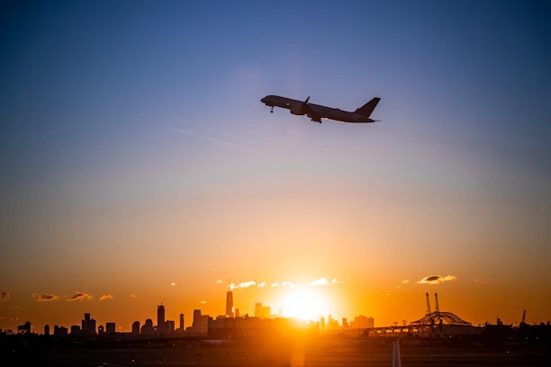 A sleek airplane soaring above clouds with a city skyline below at sunset.