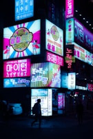 Bright neon signs illuminate a street scene at night, featuring colorful advertisements in a mix of Korean and English. Vibrant images and text cover several signs, promoting various entertainment venues and shops. People walk past, silhouetted against the glowing lights.