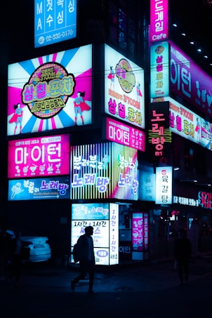 Bright neon signs illuminate a street scene at night, featuring colorful advertisements in a mix of Korean and English. Vibrant images and text cover several signs, promoting various entertainment venues and shops. People walk past, silhouetted against the glowing lights.
