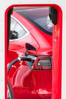 A red electric car is being charged at a charging station. The charging cable is plugged into the side of the car, and the image focuses on the rear part of the vehicle within the frame of the charging station.