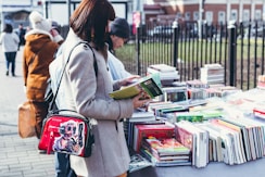 A person stands at an outdoor book market, holding and examining a book with a table full of various colorful books in front of them. They wear a light-colored coat and have a shoulder bag featuring a vibrant design. Other people are visible in the background, also browsing the books.