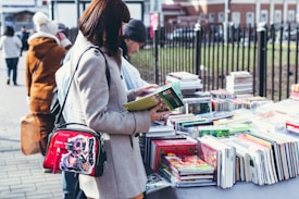 A person stands at an outdoor book market, holding and examining a book with a table full of various colorful books in front of them. They wear a light-colored coat and have a shoulder bag featuring a vibrant design. Other people are visible in the background, also browsing the books.