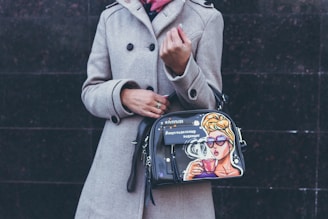 A person wearing a beige coat is holding a stylish handbag adorned with a colorful pop art design featuring a woman drinking from a cup, wearing sunglasses, and styled with a vintage look.