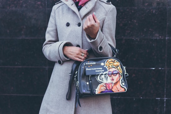 A person wearing a beige coat is holding a stylish handbag adorned with a colorful pop art design featuring a woman drinking from a cup, wearing sunglasses, and styled with a vintage look.