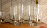 Several clear glass tumblers are neatly arranged on a wooden shelf. The background features a light-colored tile wall with a subtle geometric pattern.