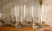 Several clear glass tumblers are neatly arranged on a wooden shelf. The background features a light-colored tile wall with a subtle geometric pattern.