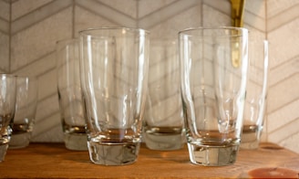 Several clear glass tumblers are neatly arranged on a wooden shelf. The background features a light-colored tile wall with a subtle geometric pattern.
