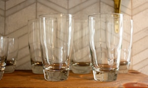 Several clear glass tumblers are neatly arranged on a wooden shelf. The background features a light-colored tile wall with a subtle geometric pattern.