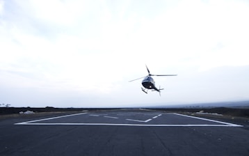 A helicopter is in the process of landing or taking off from a paved helipad situated in an open area. The sky is overcast, creating a diffused, bright light. The surrounding landscape is flat with distant trees visible along the horizon.